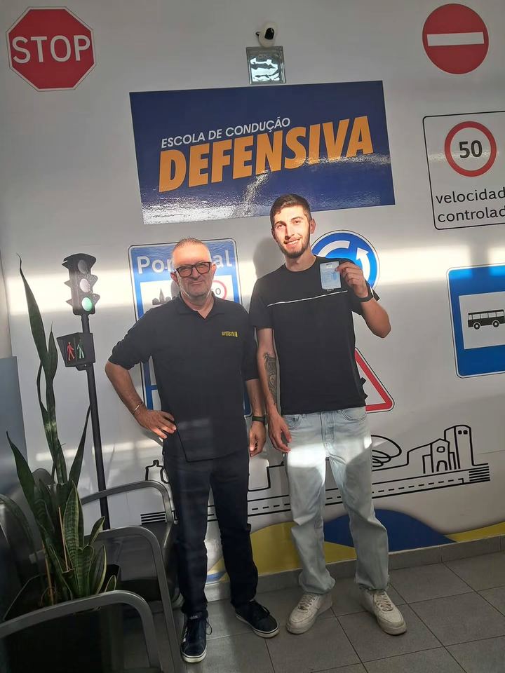Novo condutor com o instrutor, a mostrar a carta em frente à parede de sinais da escola